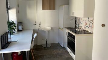 Cocina compacta con armarios blancos, horno integrado, lavamanos y zona de comedor con una silla.