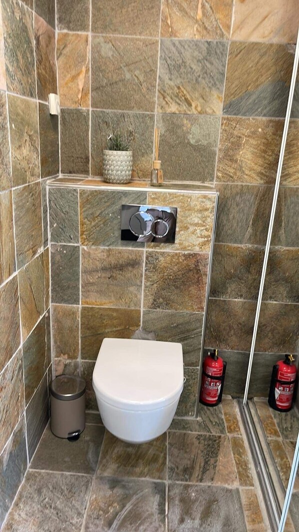 Un baño con inodoro blanco, un cesto de basura y dos extintores en la pared.