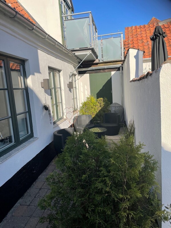 En smal innergård med en vit vägg, en liten trädgårdsdel med buskar och en balkong med ett metallräcke.