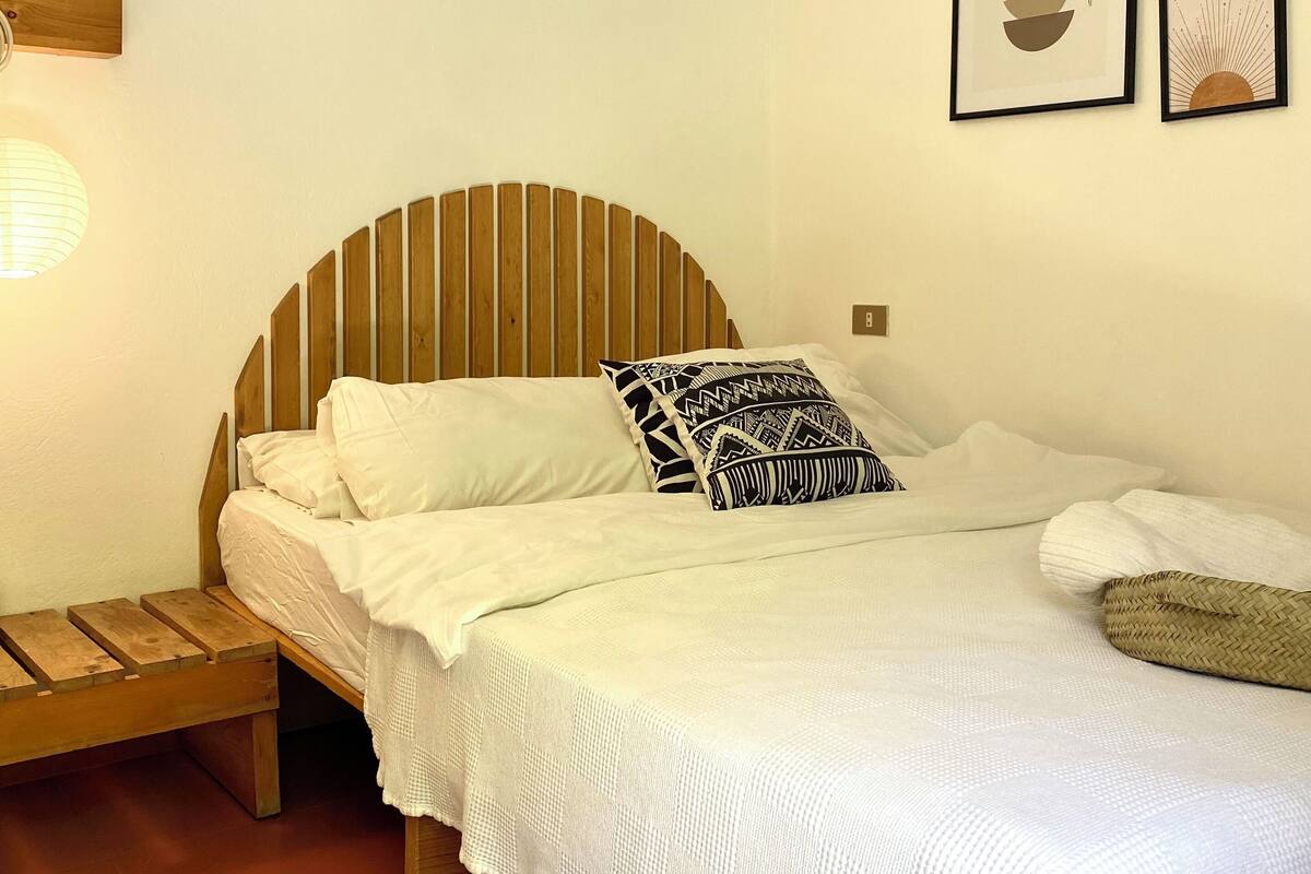 Quarto com cama de madeira, roupa de cama branca, tapete estampado e quadros nas paredes.