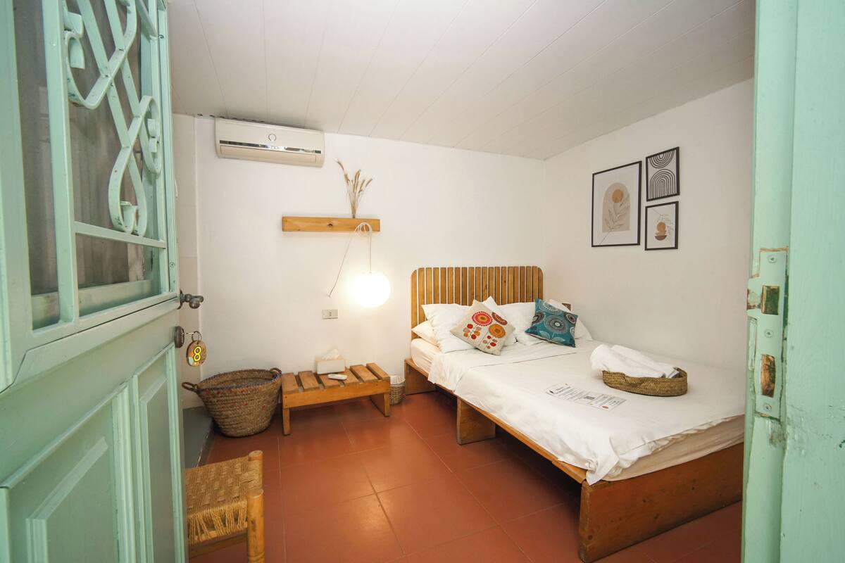 Quarto pequeno e acolhedor com uma cama de solteiro, um banco de madeira, um cesto de vime e uma prateleira na parede com uma planta.