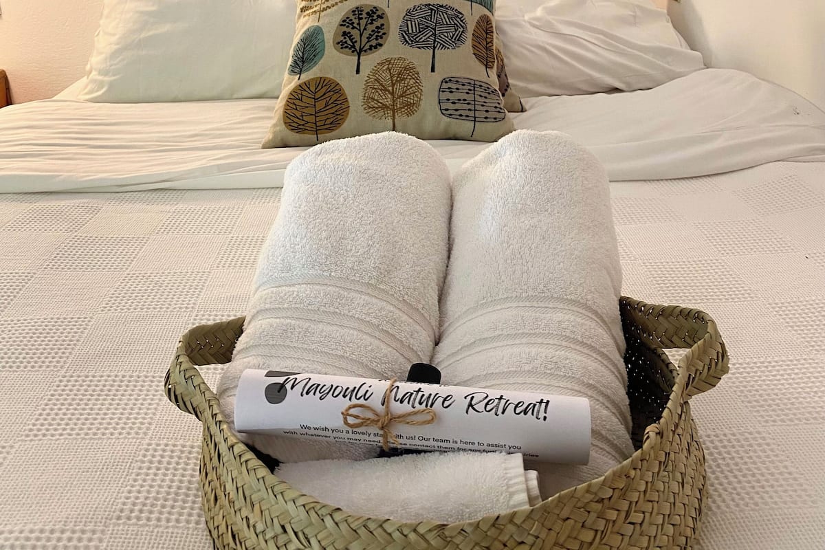 Cama bem arrumada com cabeceira de madeira, lençóis brancos e almofada decorativa com padrão natural. Uma cesta de vime com uma toalha enrolada e um convite para um refúgio está colocada sobre a cama.