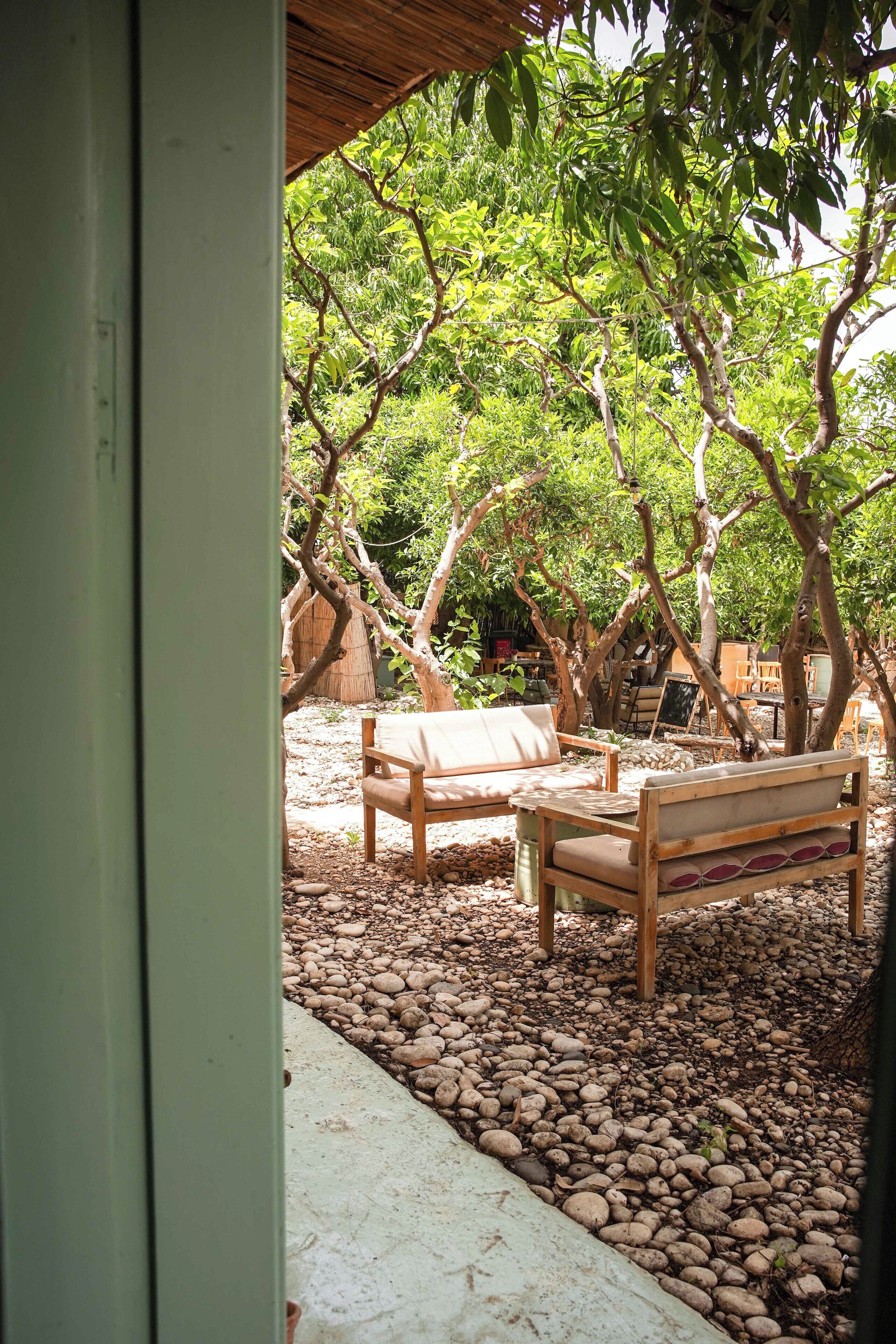 An outdoor seating area with wooden benches and a table, surrounded by trees and pebbles.