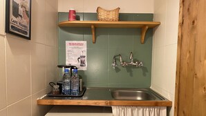 A compact kitchenette with a sink, a small fridge, a shelf with a basket and a red cup, and a poster on the wall. - Mayouli Nature Retreat (Batroun)