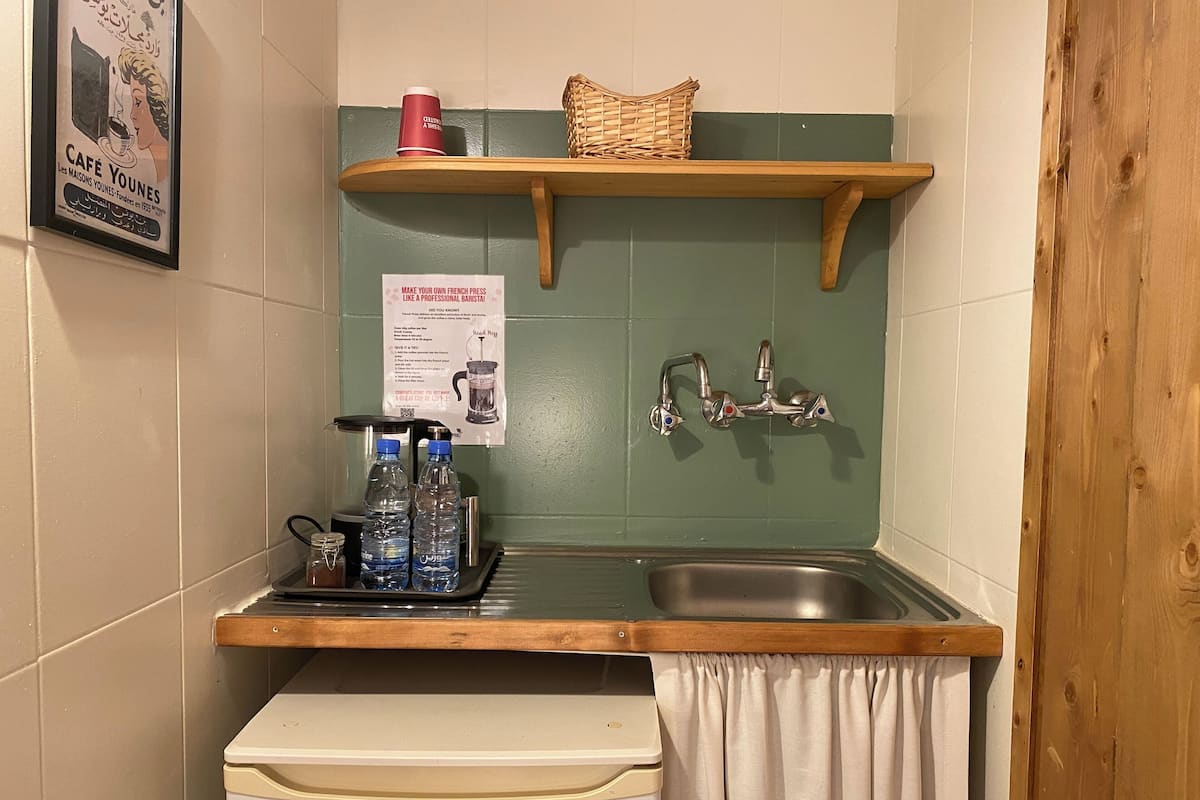 Uma kitchenette compacta com pia, uma geladeira pequena, uma prateleira com uma cesta e uma xícara vermelha, e um pôster na parede.
