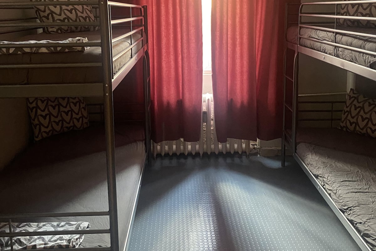A dormitory room with two bunk beds, a radiator, and a window with red curtains.