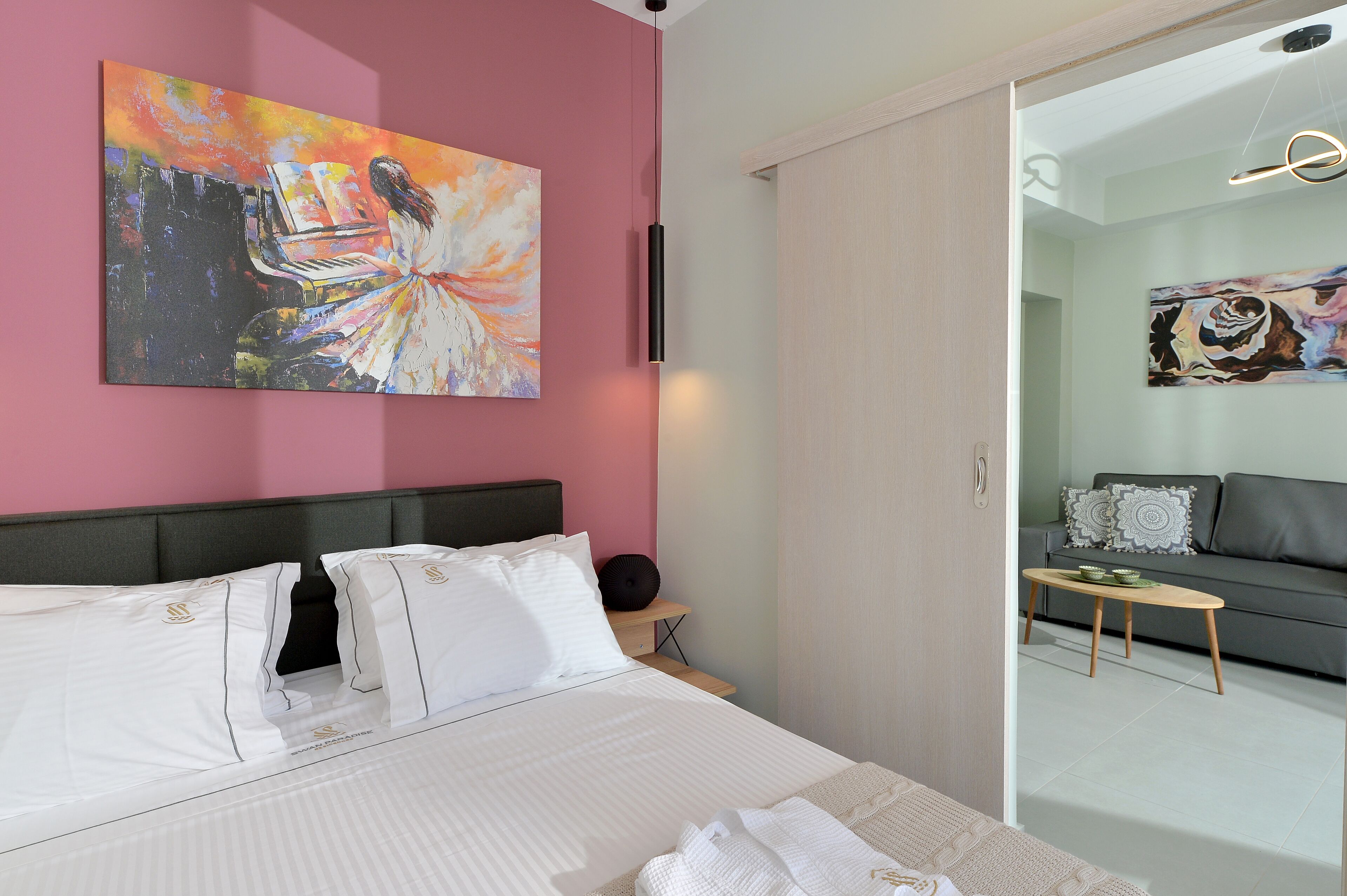A modern hotel room with a large bed, a painting on the wall, a sofa in the adjacent room, and a small table with a lamp.