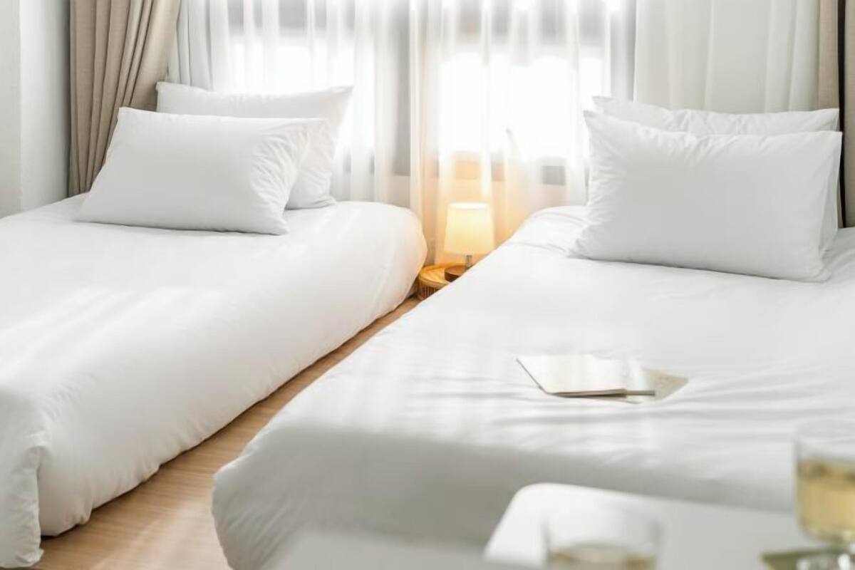 Habitación de hotel con dos camas, una mesita de noche con lámpara y una ventana con cortinas.