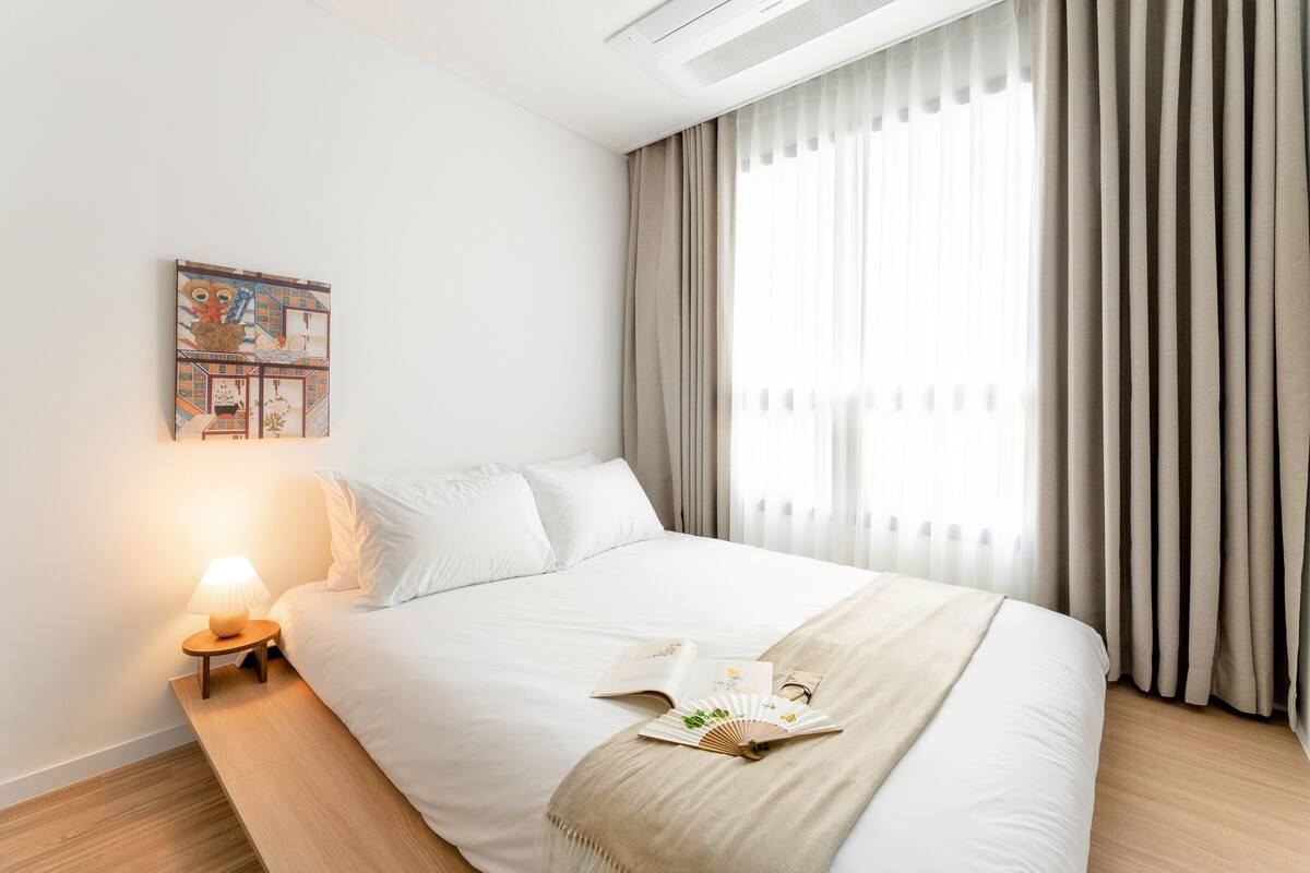 Una cama bien hecha con una colcha color beige, una lámpara de noche y un libro en la mesita.