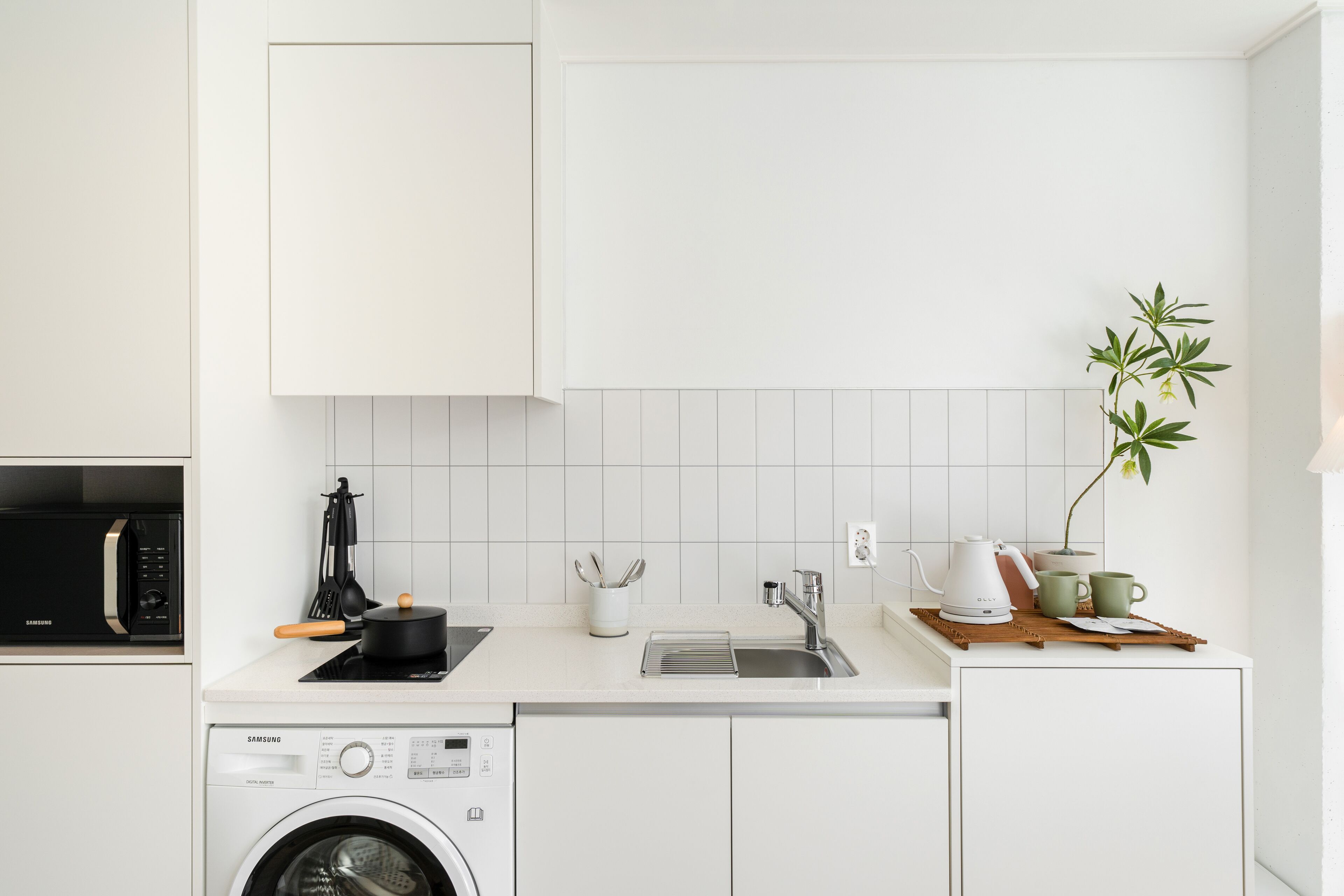 Een moderne keuken met witte kastjes, een ingebouwde magnetron, een wasmachine, een spoelbak en een kleine plant.