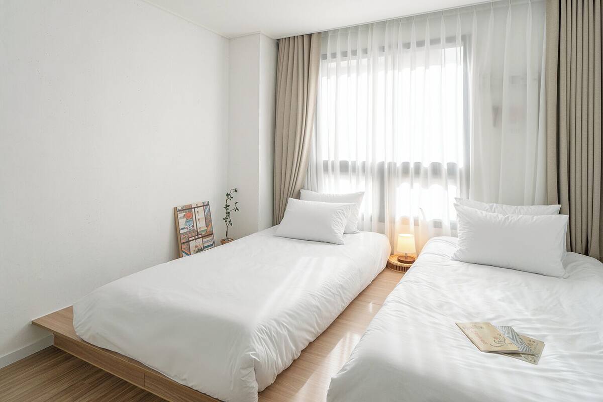 Habitación de hotel con dos camas, una mesita de noche con lámpara, una planta pequeña y un cuadro en la pared.
