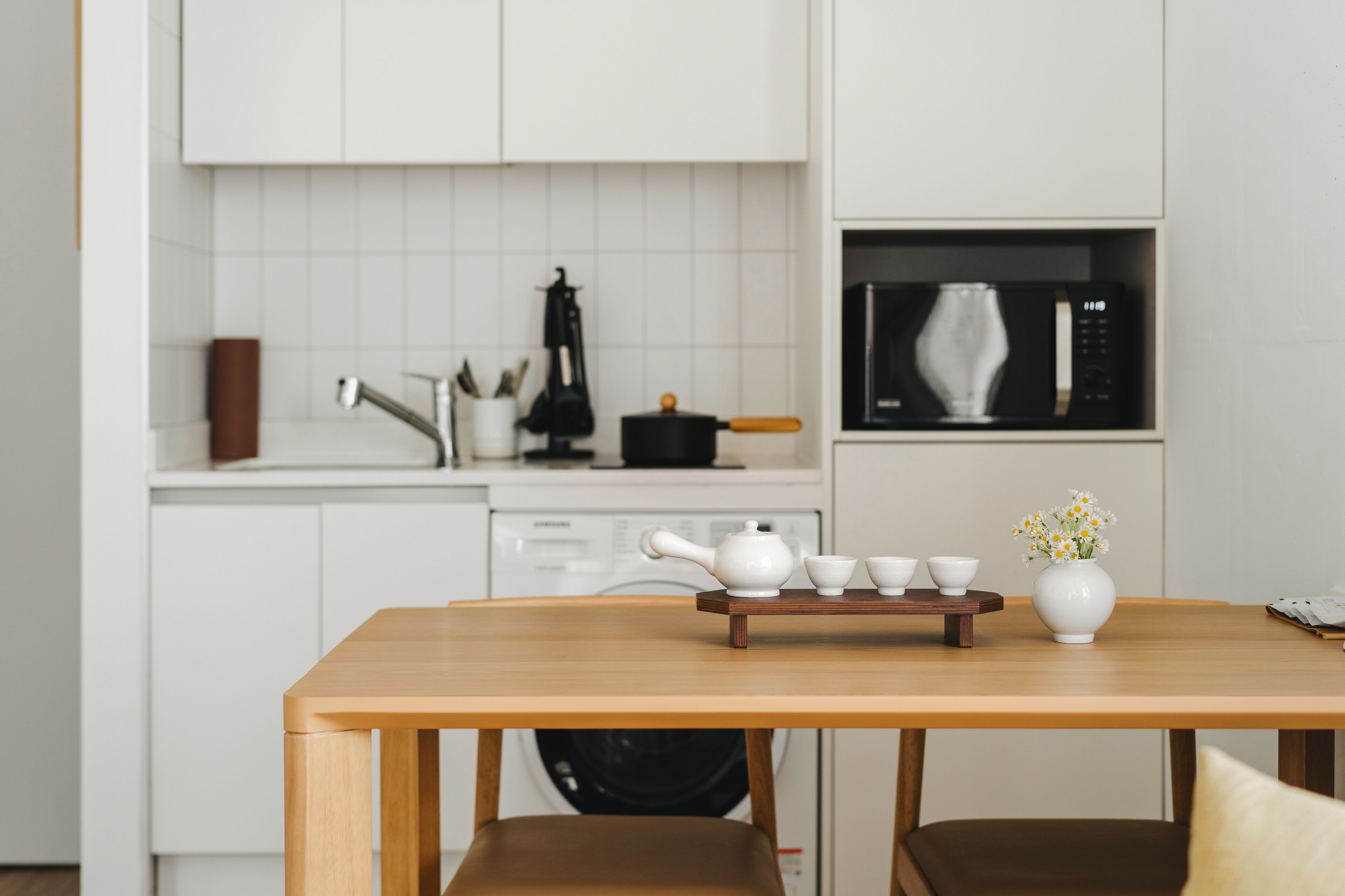 Una cocina moderna con armarios blancos, un microondas y una mesa de madera con tetera y tazas.