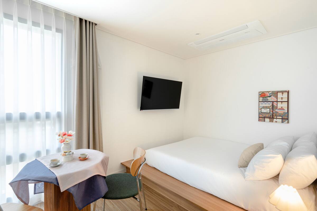 Habitación de hotel con cama, televisión montada en la pared, una mesita con silla y ventana con cortinas.