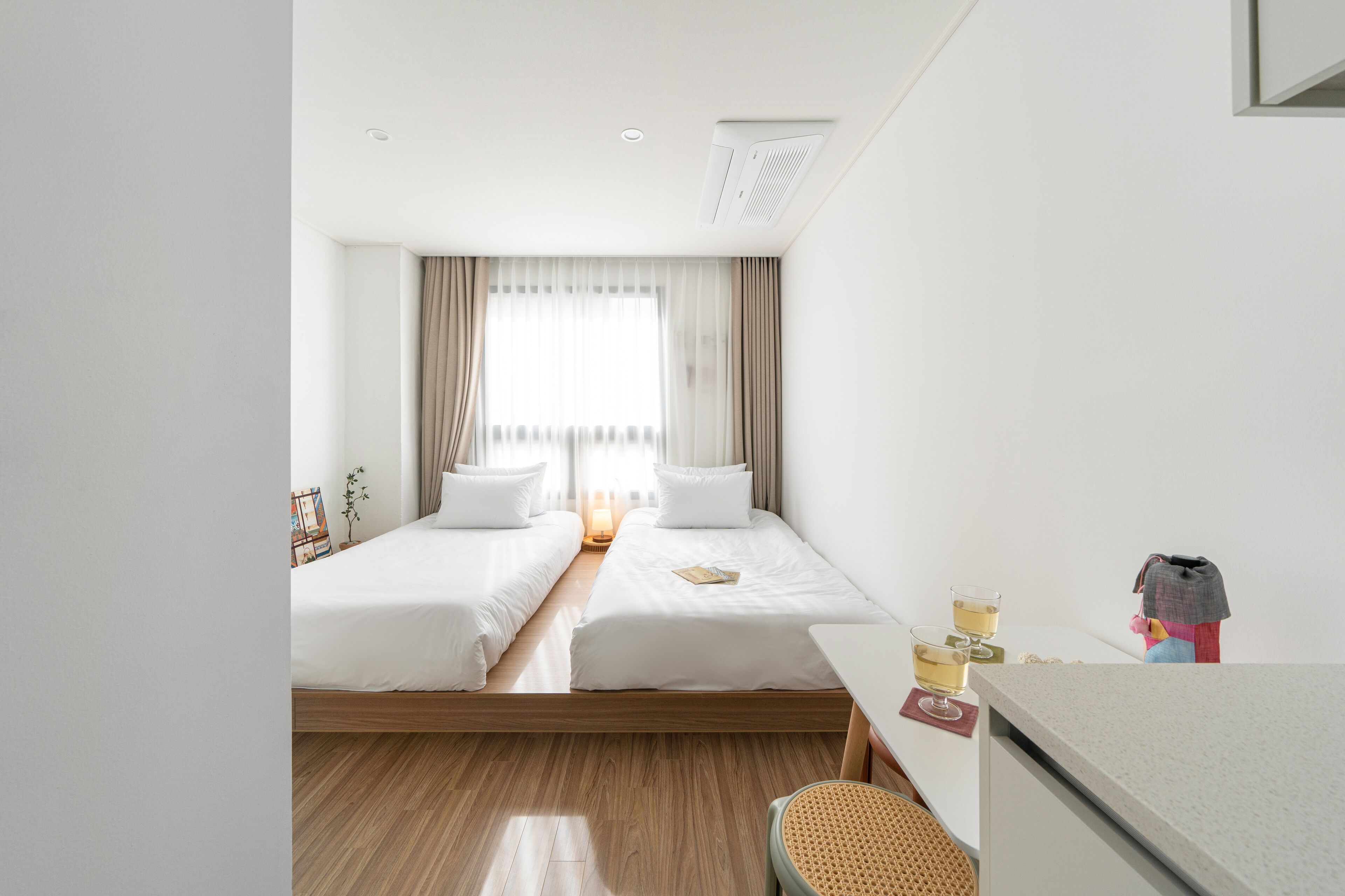 Quarto de hotel com duas camas, uma mesinha, uma cadeira e janela com cortinas.