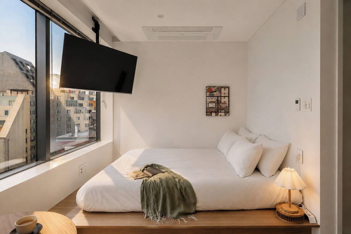 Una habitación de hotel moderna con una cama grande, un televisor montado en la pared, una mesita de noche con lámpara y vistas a edificios desde la ventana.