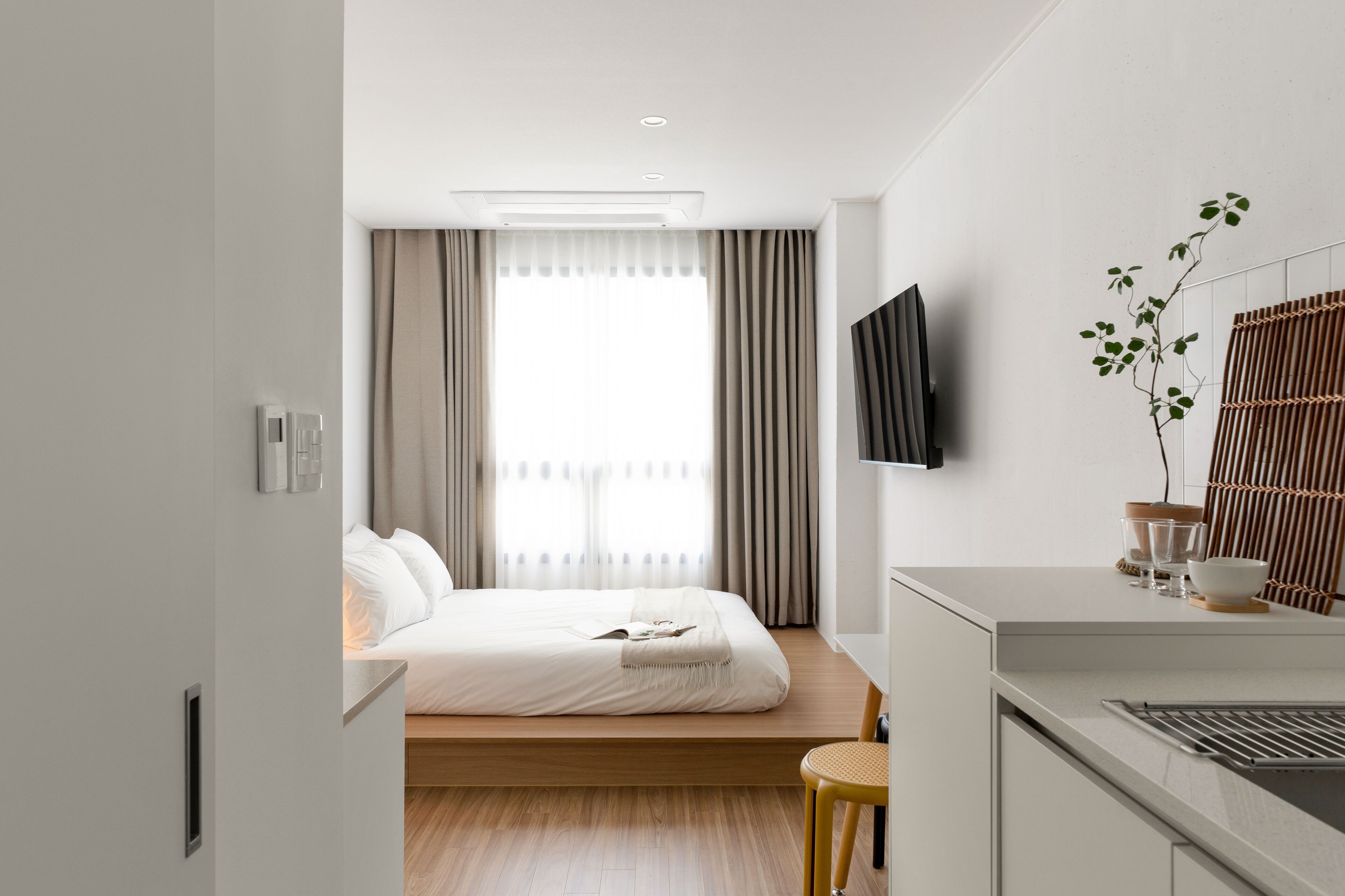 A modern hotel room with a bed, bedside table, TV, and a small stool.