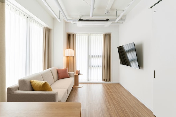 A modern hotel room with a sofa, a small side table, a flat-screen TV mounted on the wall, and large windows with curtains.