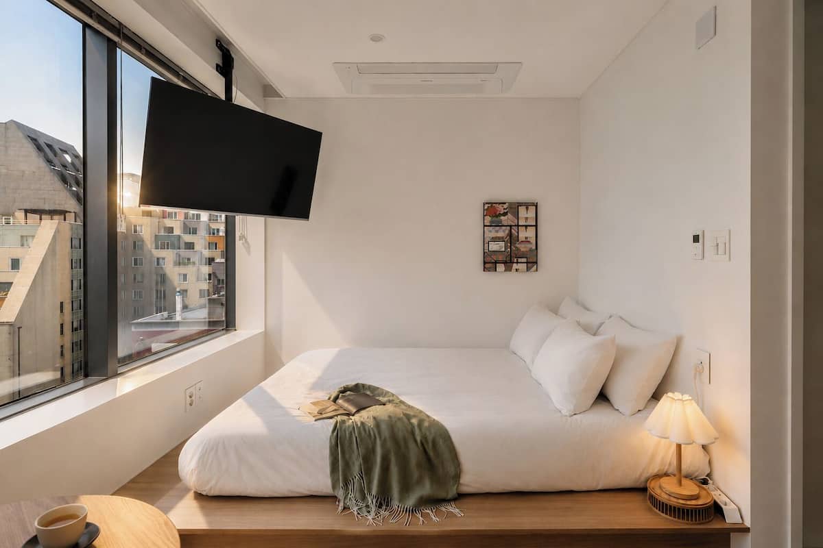 Ein modernes Hotelzimmer mit einem großen Bett, einem an der Wand montierten Fernseher, einem Nachttisch mit Lampe und Blick auf Gebäude vor dem Fenster.