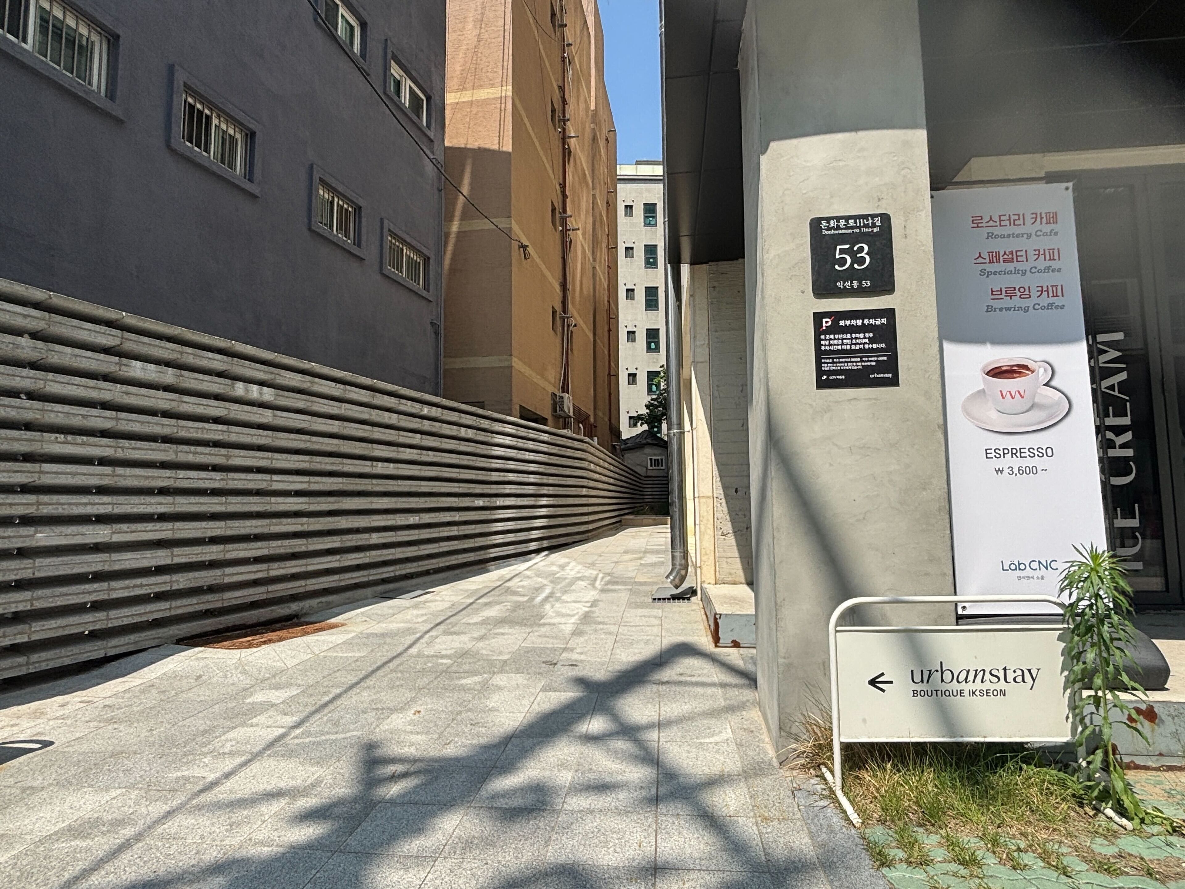 A narrow urban alleyway with a modern building on the left and a sign for 'urbanstay' boutique ikseon on the right.