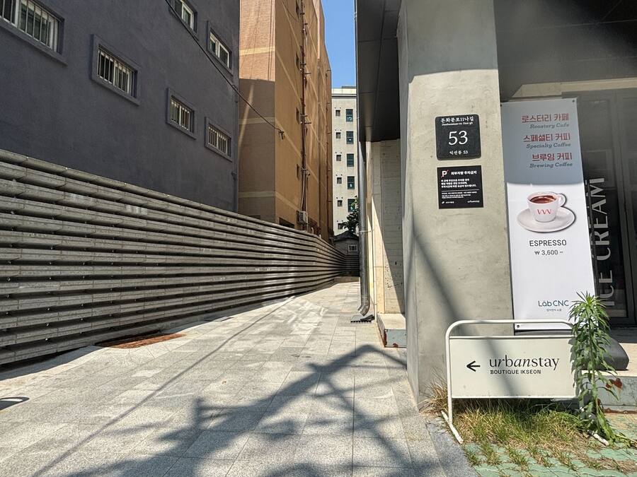 A narrow urban alleyway with a modern building on the left and a sign for 'urbanstay' boutique ikseon on the right.
