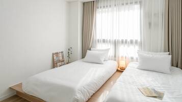 A hotel room with two beds, a bedside table with a lamp, and a window with sheer curtains.