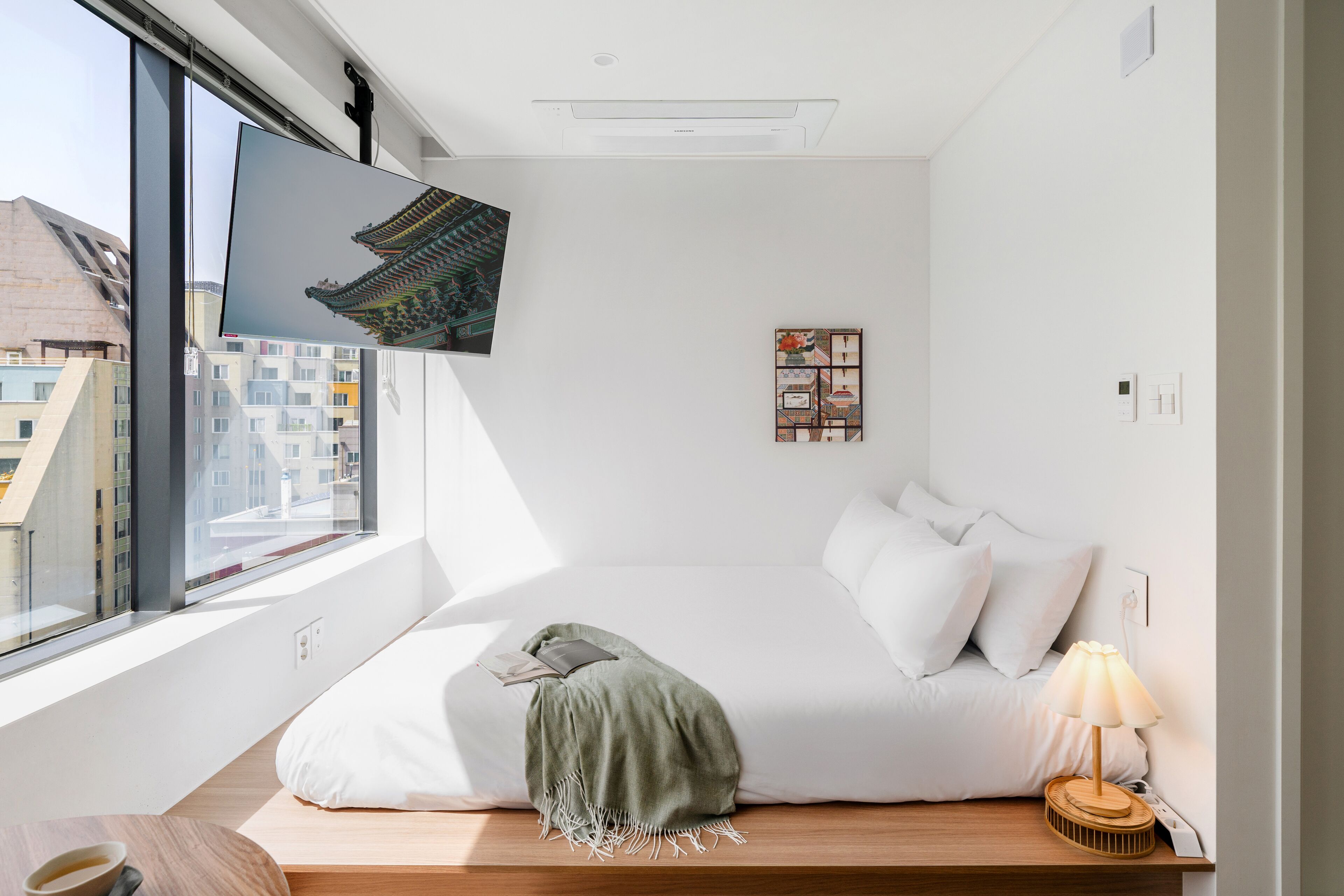 A modern bedroom with a large window, a bed, a bedside table with a lamp, and a wall-mounted TV.