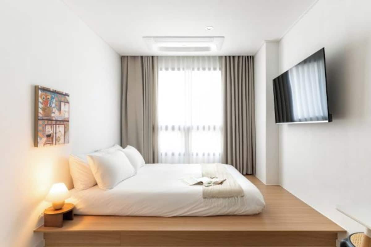 A modern hotel room with a bed, a flat-screen TV, a bedside lamp, and a window with curtains.