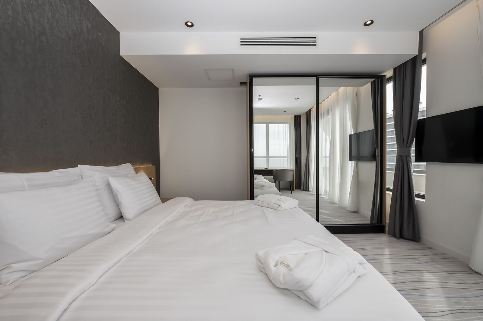 Una habitación de hotel moderna con una cama grande, un televisor de pantalla plana y vistas a la ciudad a través de un ventanal amplio.