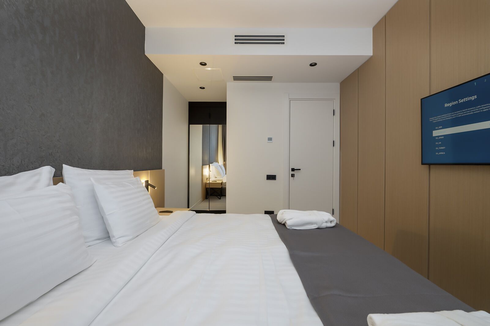 Une chambre d’hôtel avec un lit, une table de chevet et un miroir. Une porte donne accès à une salle de bain, et un écran fixé au mur affiche les paramètres de la chambre.