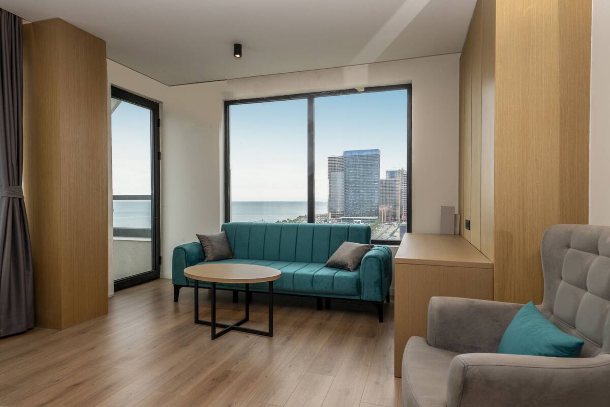 A modern room with a teal sofa, a grey armchair, and a round table. A large window offers a view of the city skyline and water.