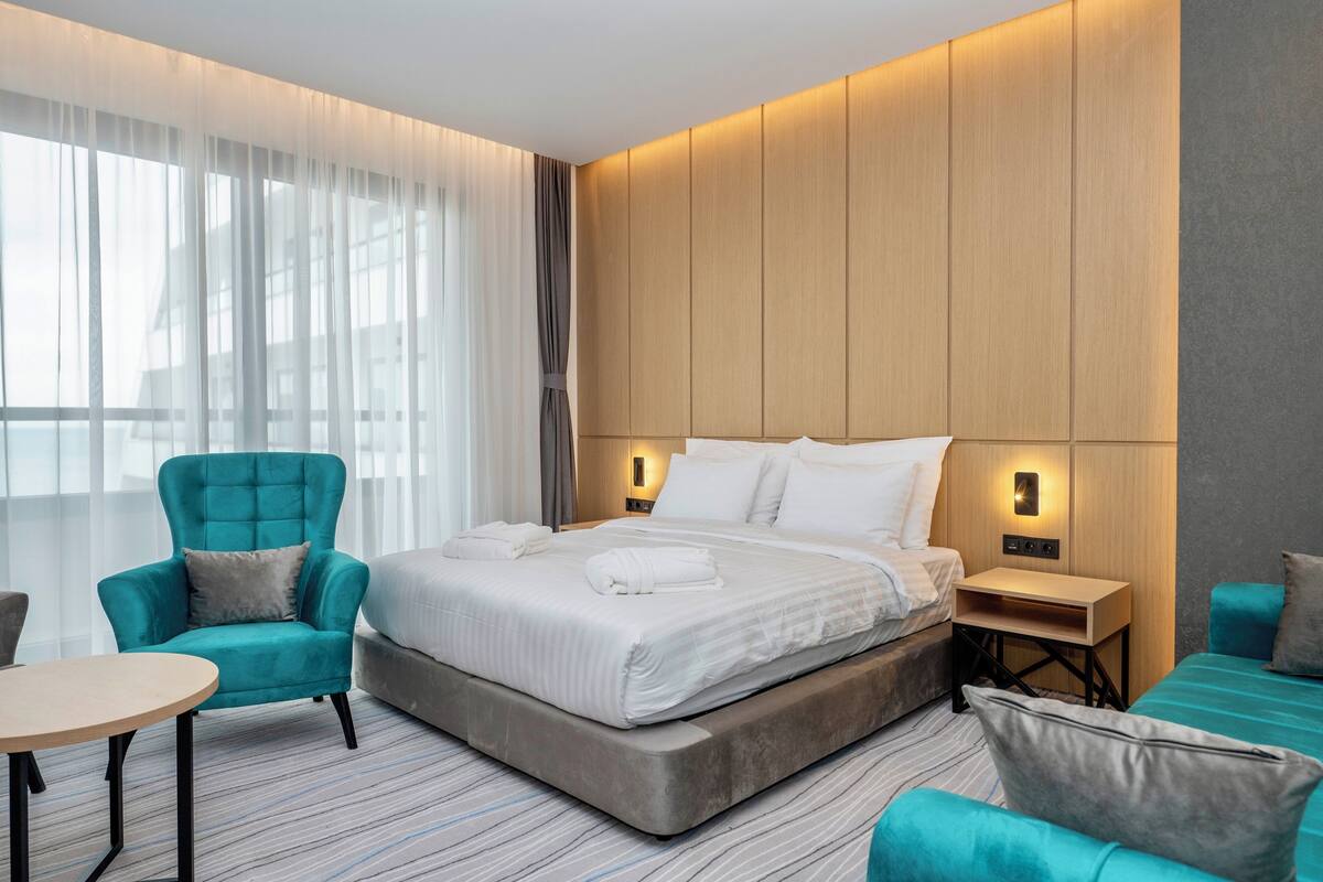 A modern hotel room with a large bed, a seating area with a chair and a table, and a built-in desk with a lamp.