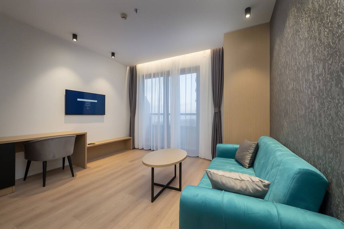 A modern living room with a teal sofa, a round coffee table, a desk, and a TV mounted on the wall.