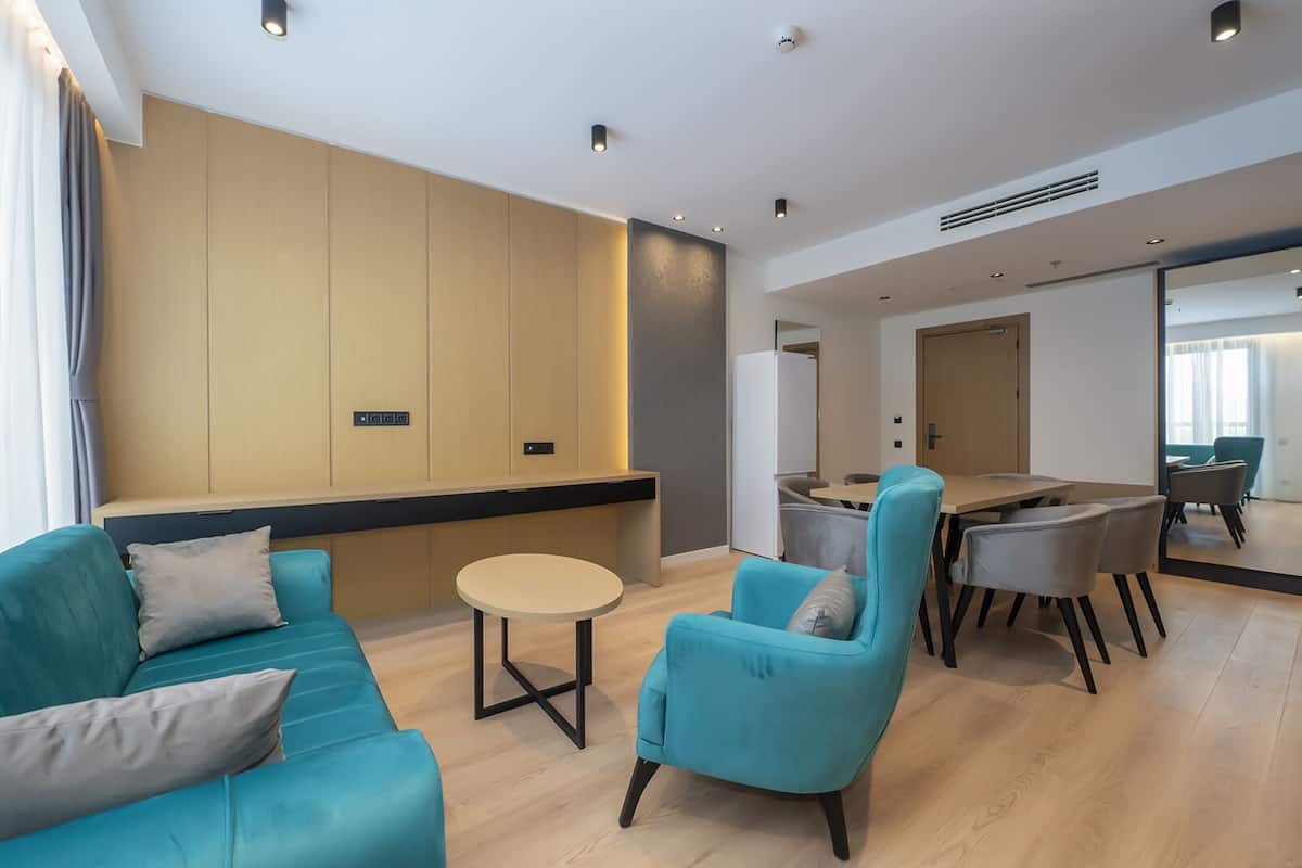 A modern hotel room with teal sofas, a round table, and a dining area with chairs.