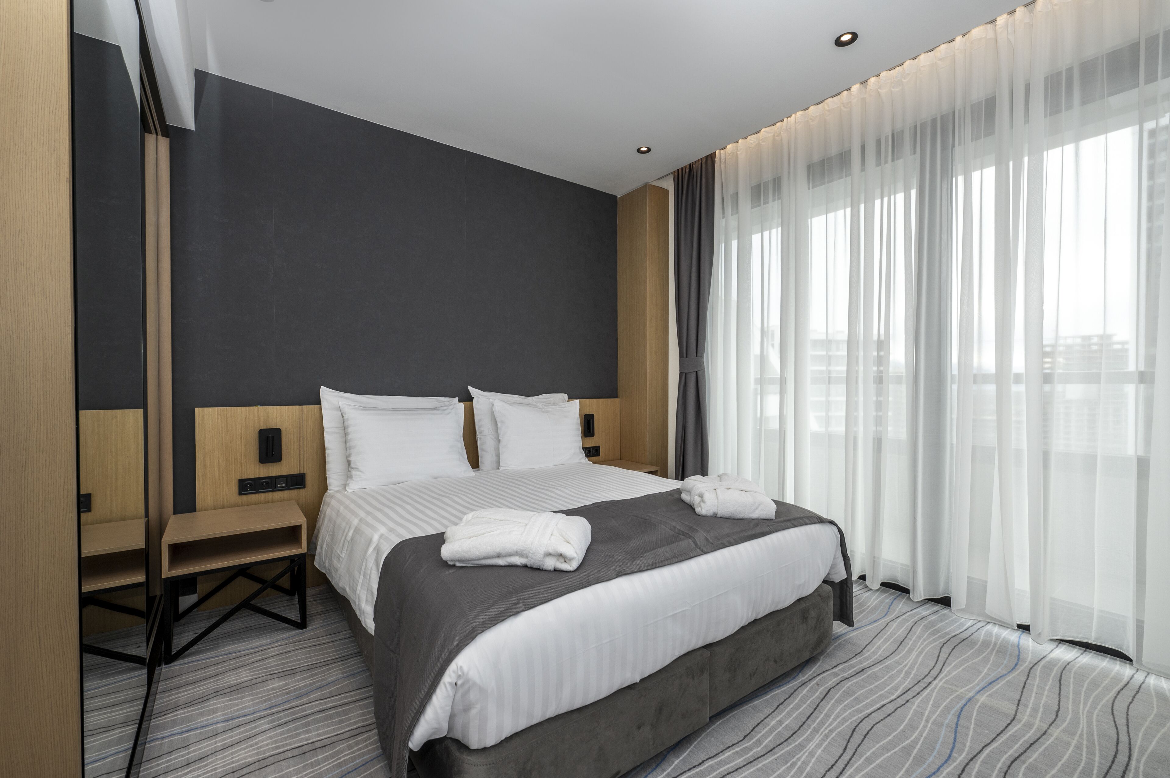 A modern hotel room with a large bed, a wooden nightstand, a small table, and a large window with sheer curtains.