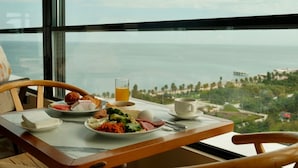 Una mesa de desayuno con alimentos y un vaso de jugo de naranja, con vistas a un paisaje con playa y palmeras.