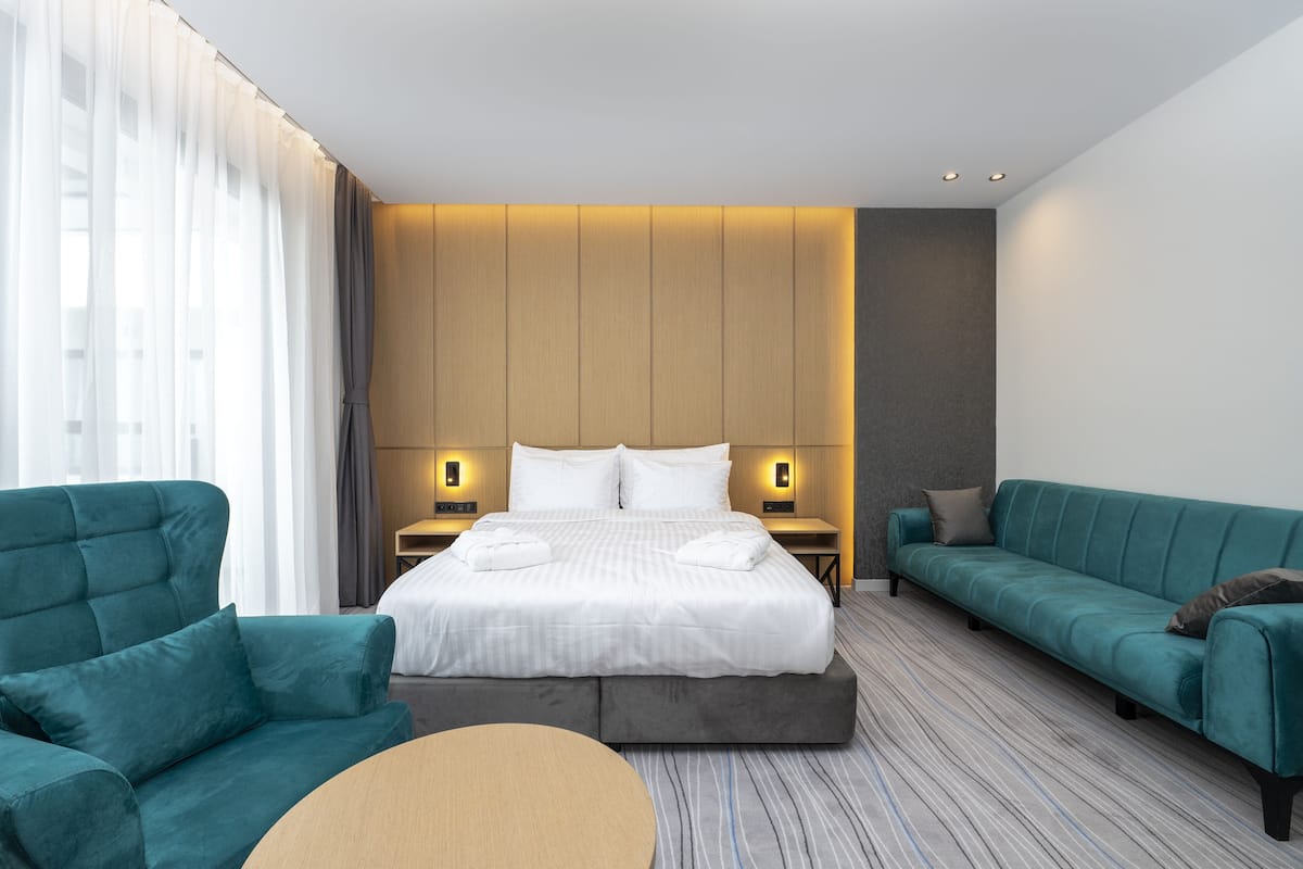 A modern hotel room with a large bed, two green sofas, a round table, and a view of the city skyline.