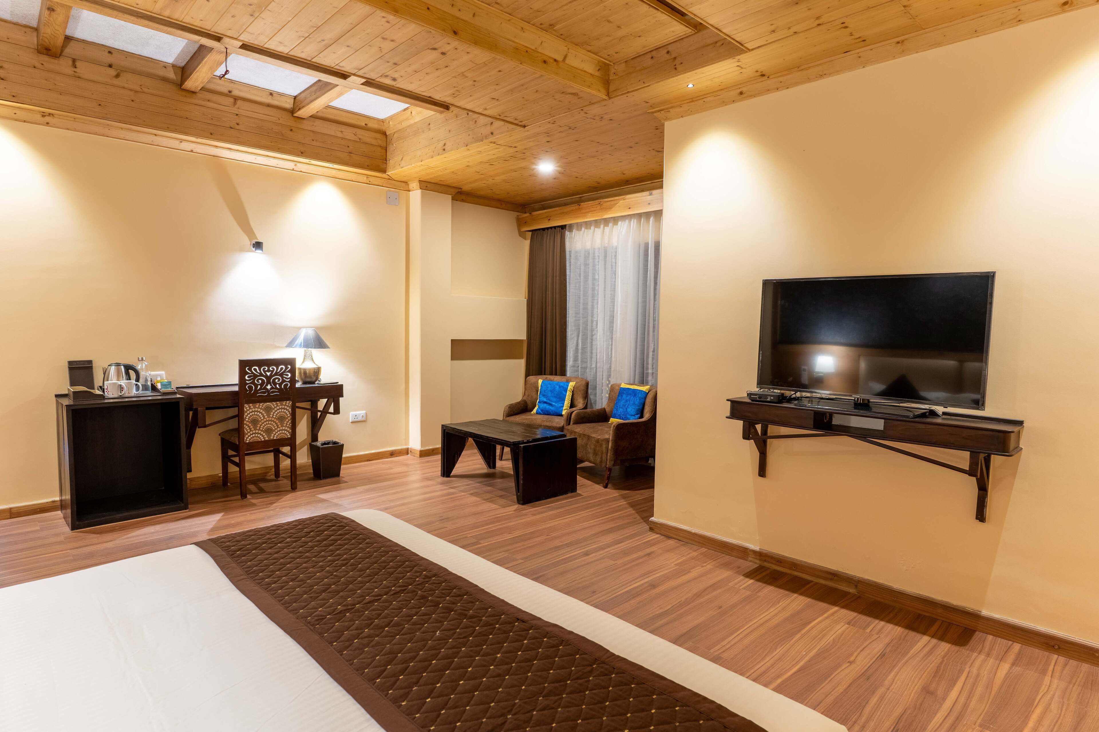 A cozy living room with a wooden ceiling, a flat-screen TV mounted on the wall, a coffee table, two armchairs, a small side table, and a rug.