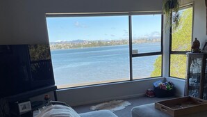 A living room with a large window offering a view of a body of water and distant land. - Unique property overlooking estuary to city on the beautiful Daisy Hardwick walk (Tauranga)