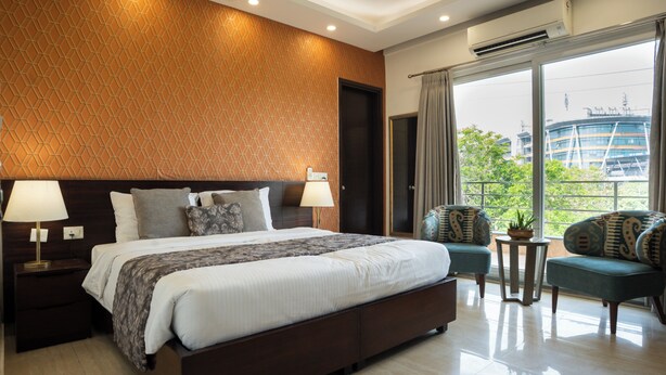 Une chambre d’hôtel moderne avec un grand lit, deux tables de chevet avec des lampes, une petite table d’appoint avec une plante, et un balcon offrant une vue sur les bâtiments et les arbres.