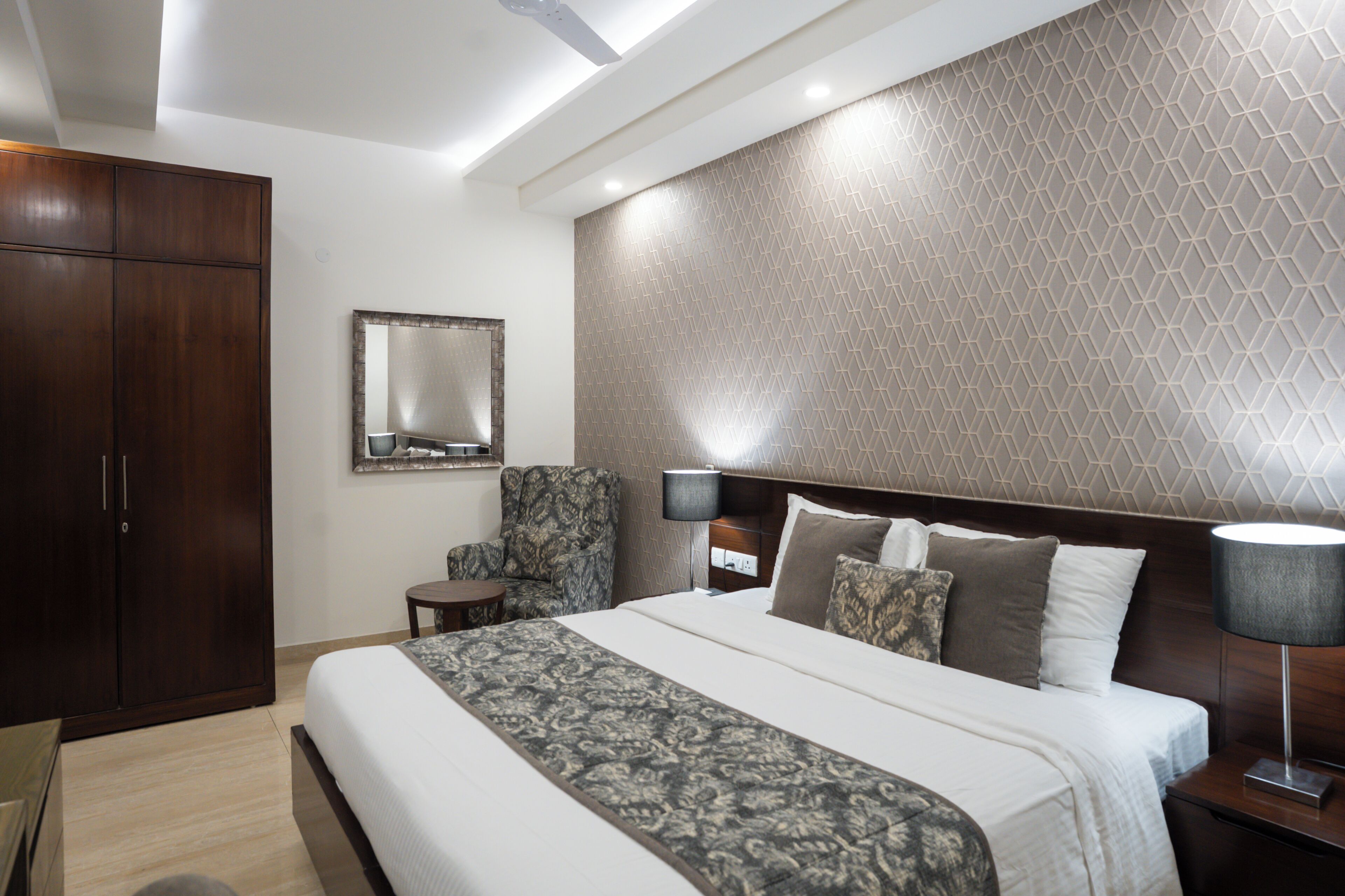 Une chambre d’hôtel avec un grand lit, une armoire, une chaise, une table de chevet et une tête de lit ornée d’un motif géométrique.