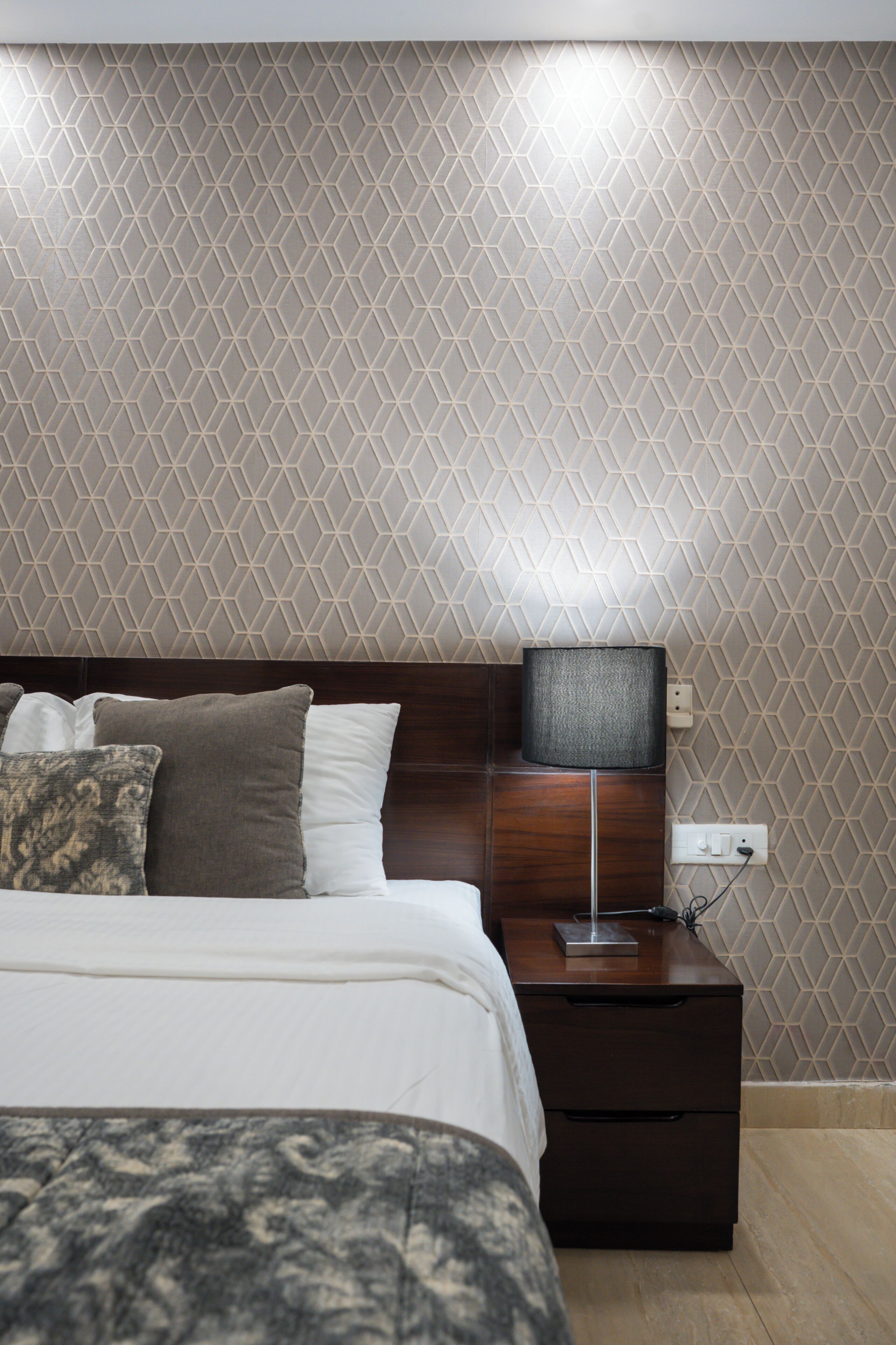 Een net opgemaakt bed met kussens, een nachtkastje met een lamp, en een muur met een geometrisch patroon.