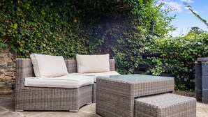 Een rieten loungeset met kussens en een salontafel met glazen blad in een patio met een stenen muur en weelderige beplanting op de achtergrond.