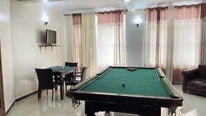 Una mesa de billar con superficie de fieltro verde, un televisor montado en la pared y una mesa de comedor con sillas.