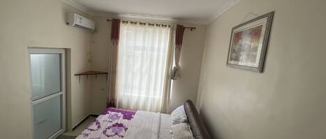 Un dormitorio con cama, mesita de noche, armario, ventana con cortinas y aire acondicionado instalado en la pared.
