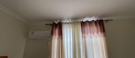 Une pièce avec une fenêtre recouverte de rideaux à motifs, un climatiseur fixé au mur et un coin de mur visible.