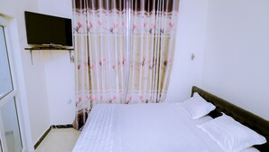 Un dormitorio con cama, ropa de cama blanca, televisor montado en la pared y cortina con estampado floral.