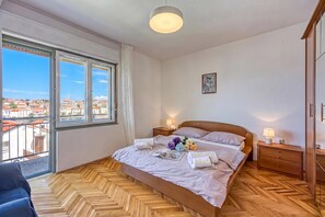 Een slaapkamer met een groot bed, houten parketvloer, stadszicht door raamhoog plafond, en een klein nachtkastje met een lamp.