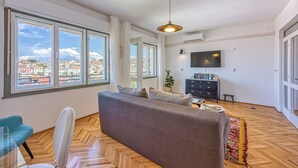 A spacious living area with a sofa, a television, and large windows offering a city view.