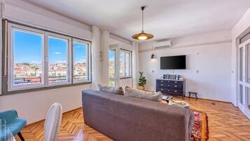 Un ampio soggiorno con divano, televisore e grandi finestre che offrono vista sugli edifici.