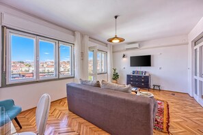 A spacious living room with a sofa, a television, and large windows offering a view of buildings. - Enolabee Harbour View Apartment (Mali Losinj)